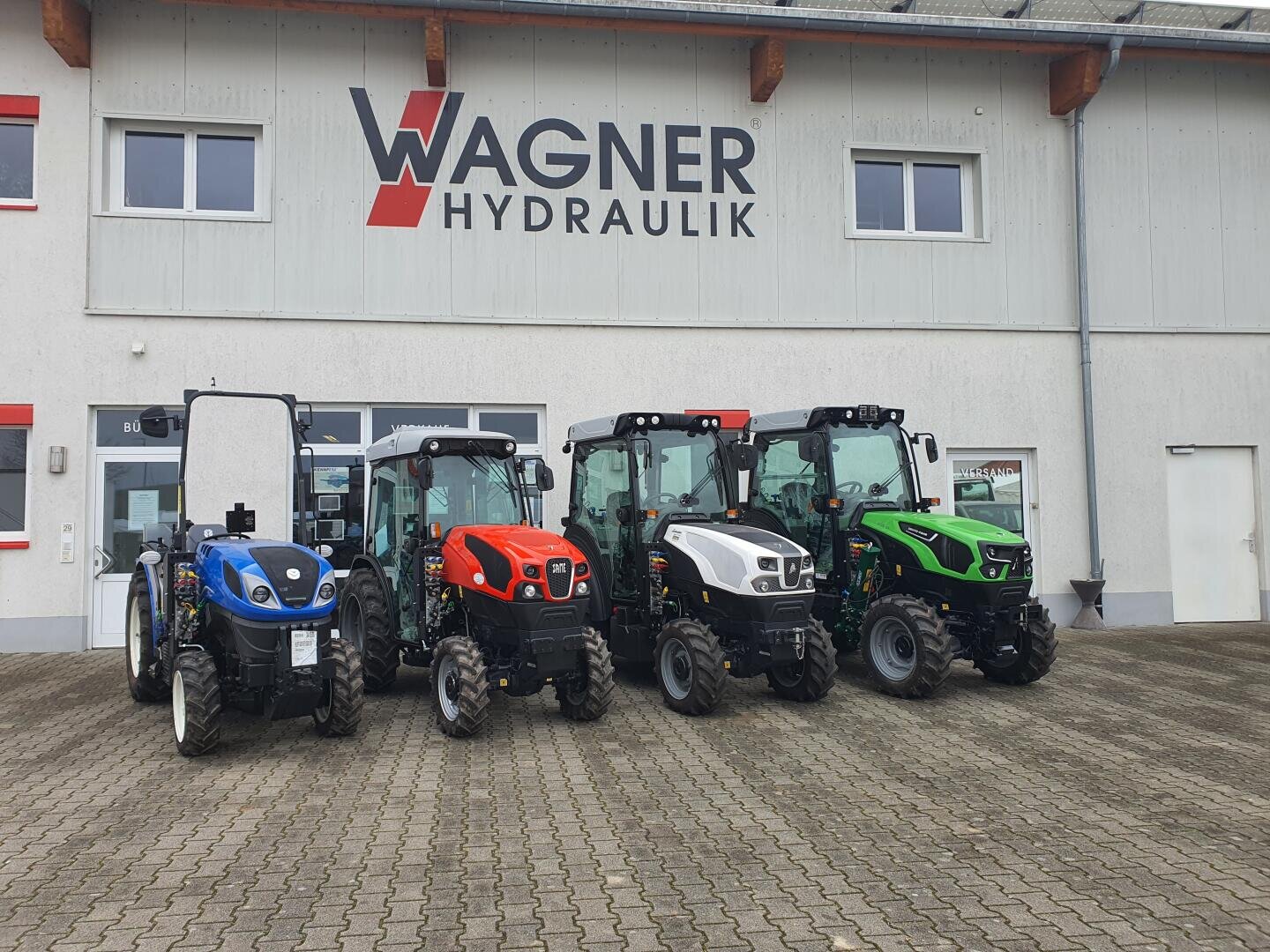Four small tractors in the colors blue, red, white and green are parked in front of a building with a sign that reads Wagner Hydraulik. The area is paved and the building has several windows.