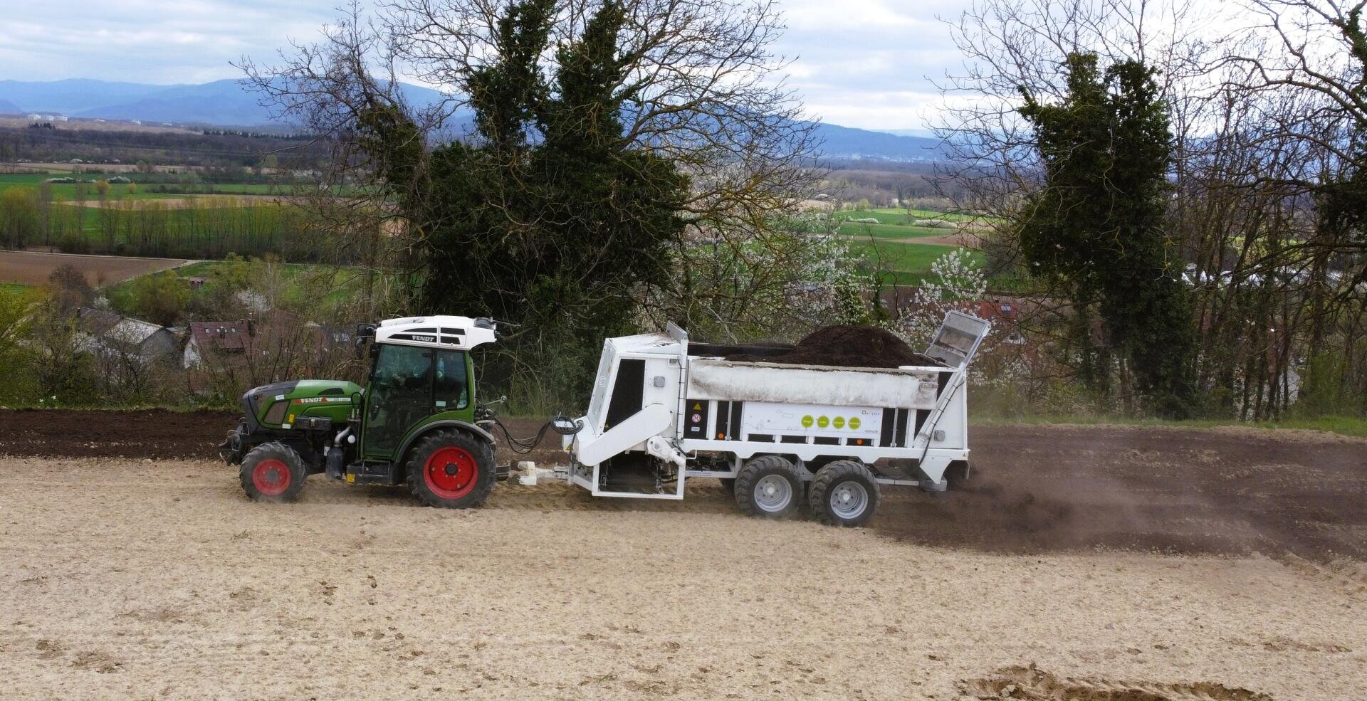 Ein grüner Traktor zieht einen weißen Düngerstreuer, der dunkles organisches Material auf ein Feld ausbringt, mit Bäumen und einer ländlichen Landschaft im Hintergrund unter einem bewölkten Himmel.