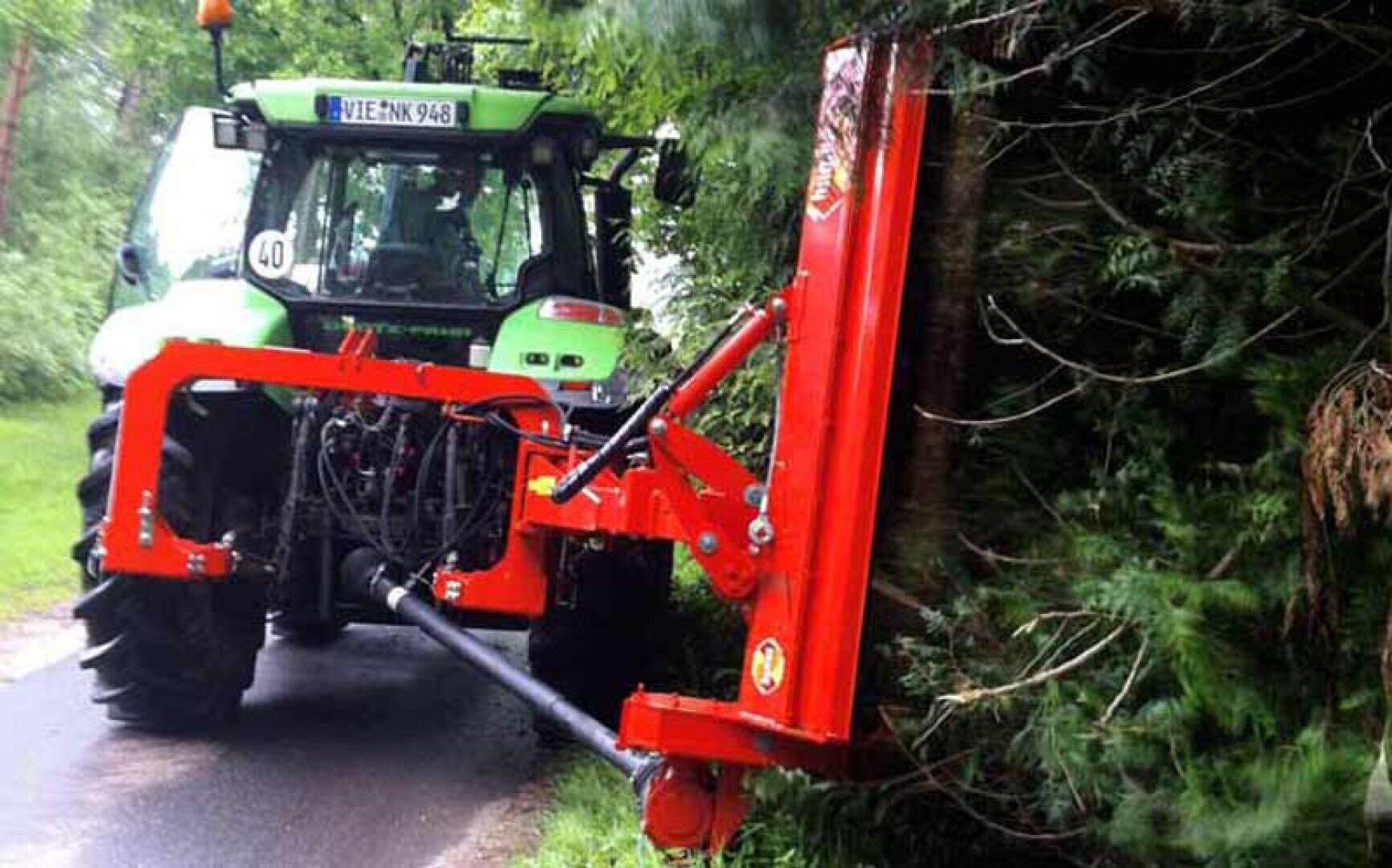 A tractor with a red mechanical arm attachment cuts overgrown branches at the side of a country road, surrounded by greenery.