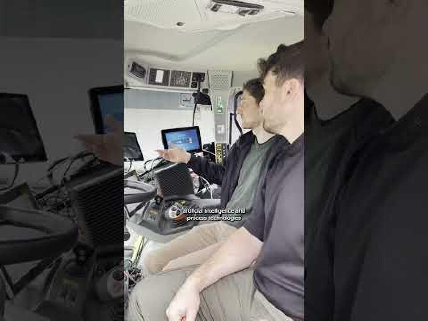 Two people are sitting in a vehicle equipped with electronic devices and screens. One person gestures towards a monitor displaying information. The scene looks technical, the caption reads "Artificial intelligence and process technologies".