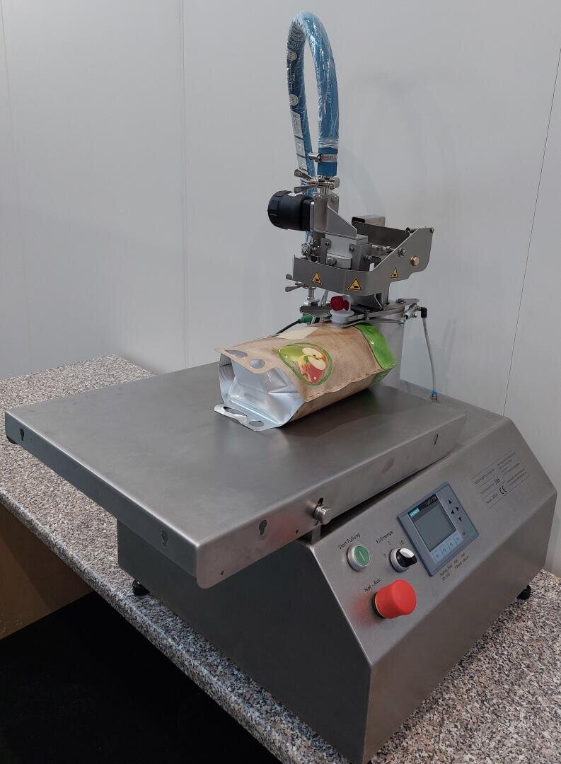 Eine Verpackungsmaschine versiegelt einen braunen und grünen Beutel mit Lebensmitteln auf einer Plattform aus rostfreiem Stahl, auf deren Vorderseite Tasten und ein Bildschirm zu sehen sind. Die Maschine steht auf einer gesprenkelten Arbeitsplatte.