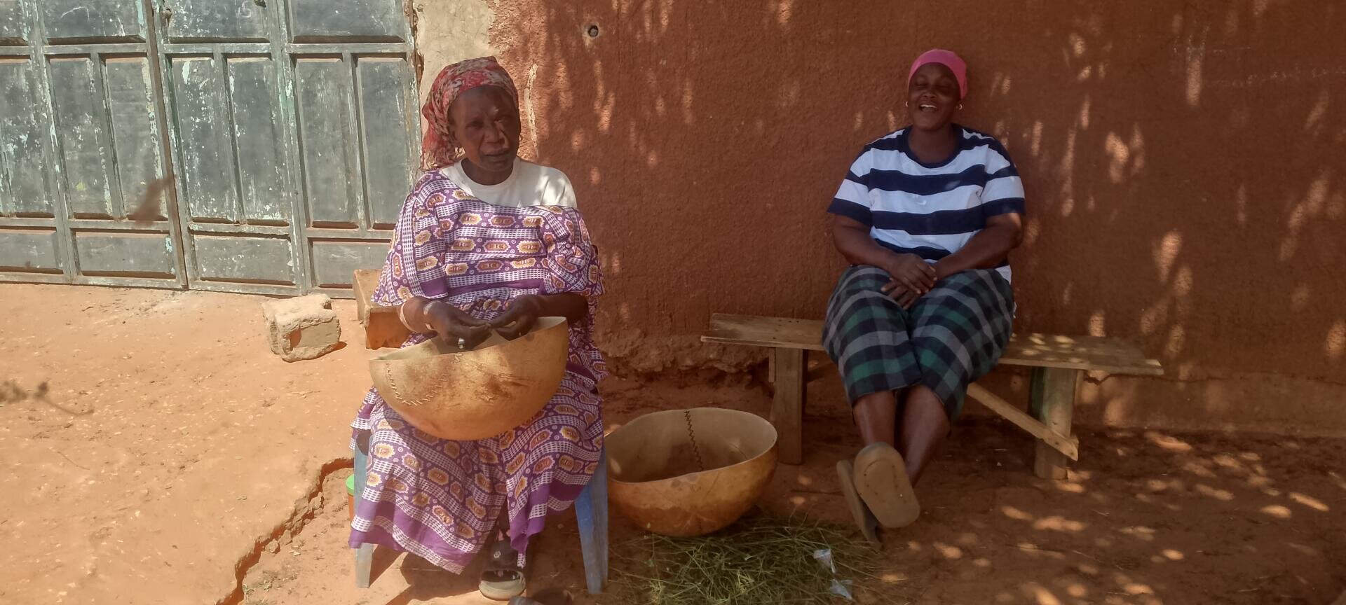 Two women sit outside in front of a reddish wall; one wears a patterned dress and holds a large calabash bowl, the other wears a striped shirt and skirt and sits smiling on a wooden bench. The sunlight casts shadows on the floor.