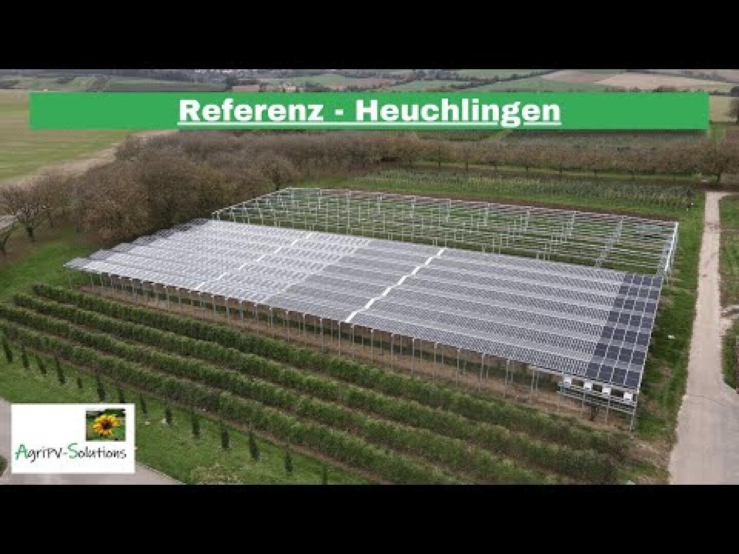 Aerial view of a large solar installation in a rural area, surrounded by fields and trees. The text Reference - Heuchlingen appears at the top, and a company logo can be seen in the bottom left-hand corner.