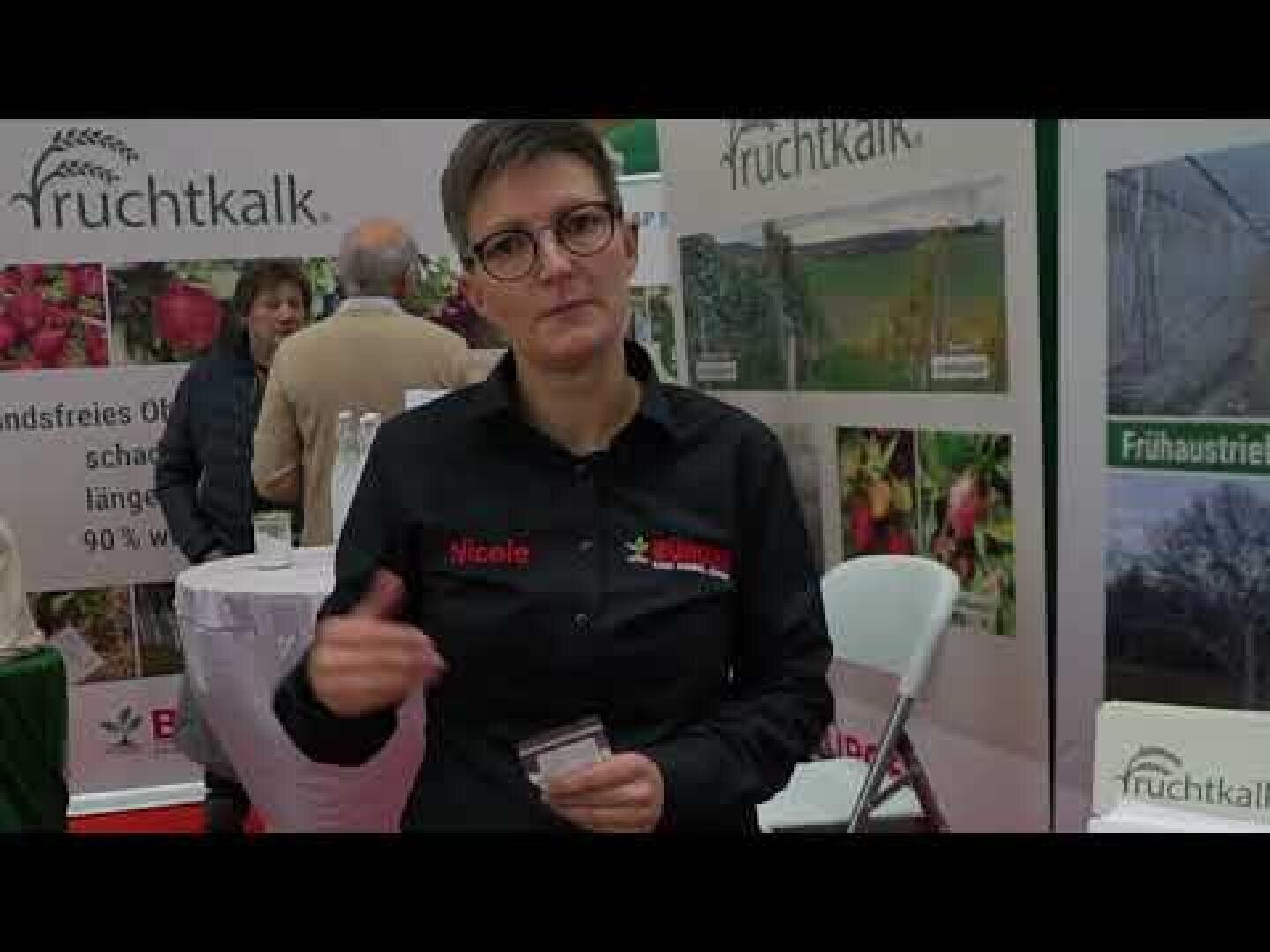 Eine Person mit kurzen Haaren und Brille steht an einem Messestand und hält eine Karte in der Hand. Hinter ihr sind Banner mit dem Wort fruchtkalk und Bildern von Früchten und Pflanzen zu sehen. Im Hintergrund sind weitere Personen zu sehen.