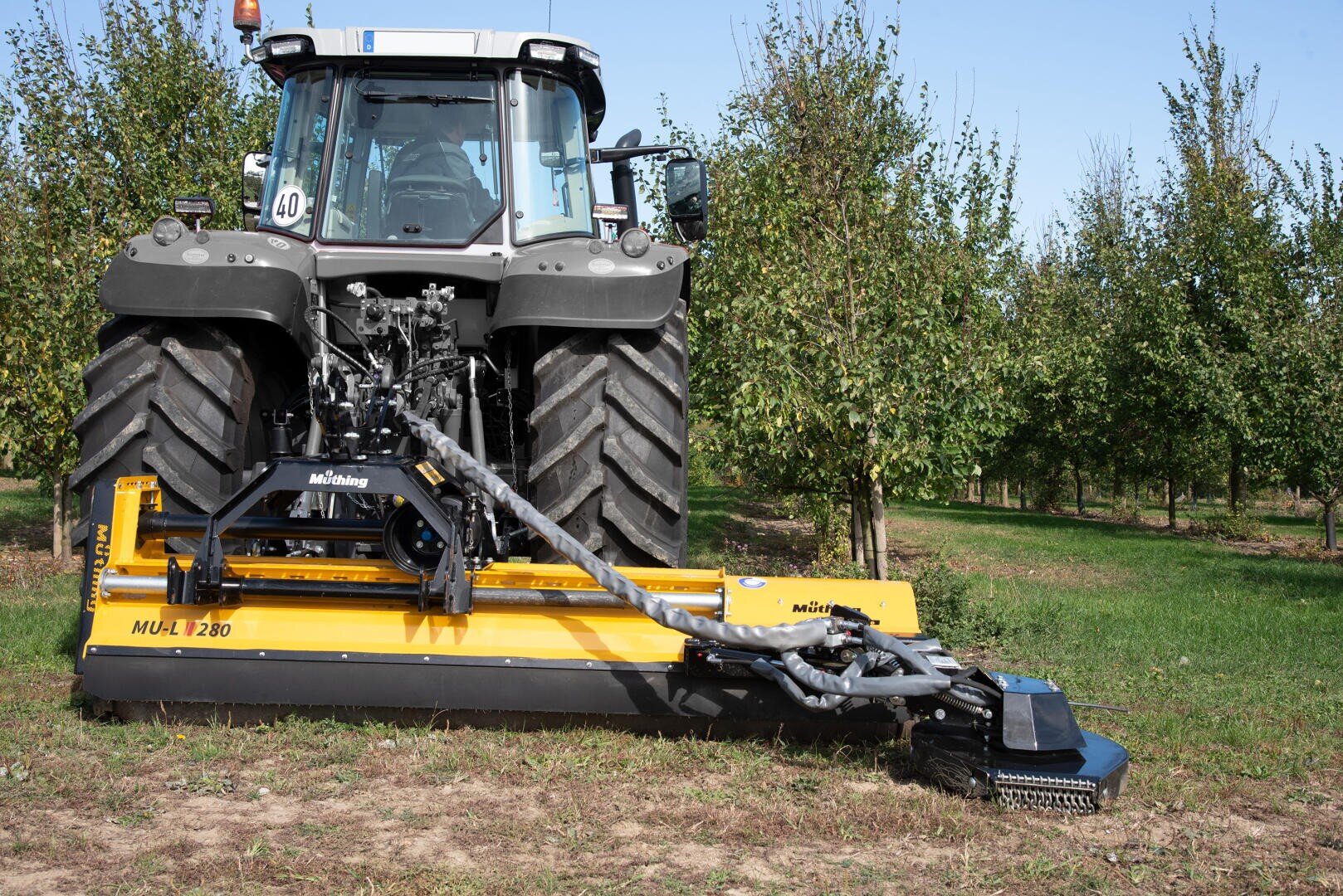 Ein großer schwarzer Traktor mit einem gelben MUL 1280-Mulchgerät ist auf einer Wiese in einem Obstgarten geparkt, mit grünen Baumreihen im Hintergrund unter einem klaren blauen Himmel.