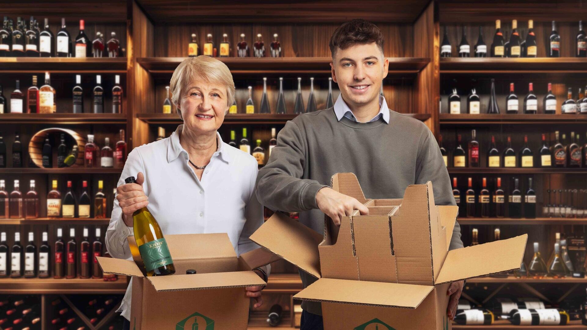 Zwei Personen, eine ältere und eine jüngere, stehen in einer Weinhandlung und verpacken lächelnd Weinflaschen in Pappkartons. Im Hintergrund sind Regale zu sehen, die mit verschiedenen Weinflaschen gefüllt sind.
