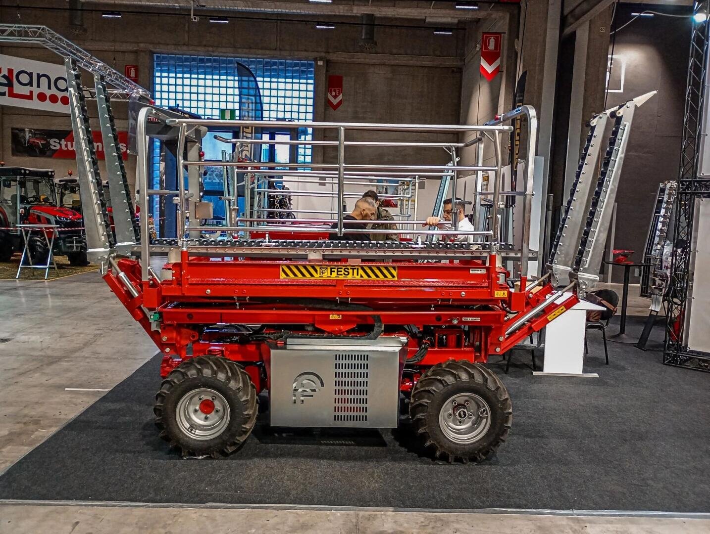 A red agricultural scissor lift machine with extended metal arms displayed indoors at an expo, with people and exhibition booths in the background.