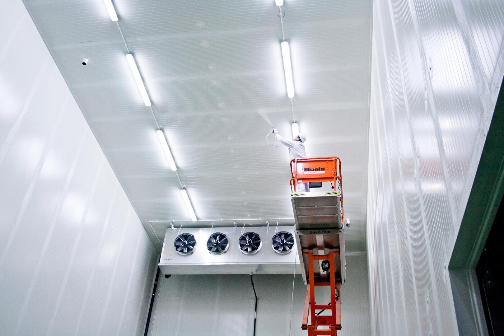 A person in a bright orange elevator works near the ceiling of a large, white industrial room with fluorescent lights and a row of large fans on one wall.
