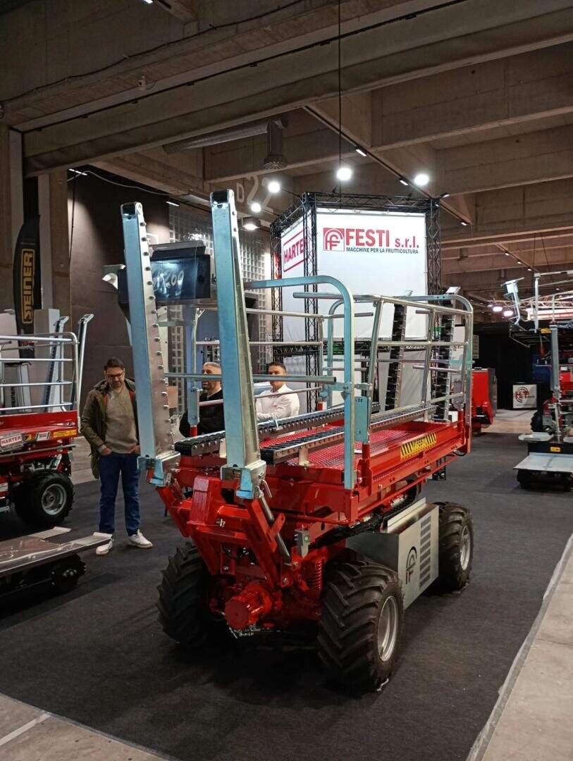 A red, heavy-duty scissor lift with large black tires is displayed indoors at an industrial expo. Two men stand nearby, and a FESTI S.r.l. company sign is visible in the background.