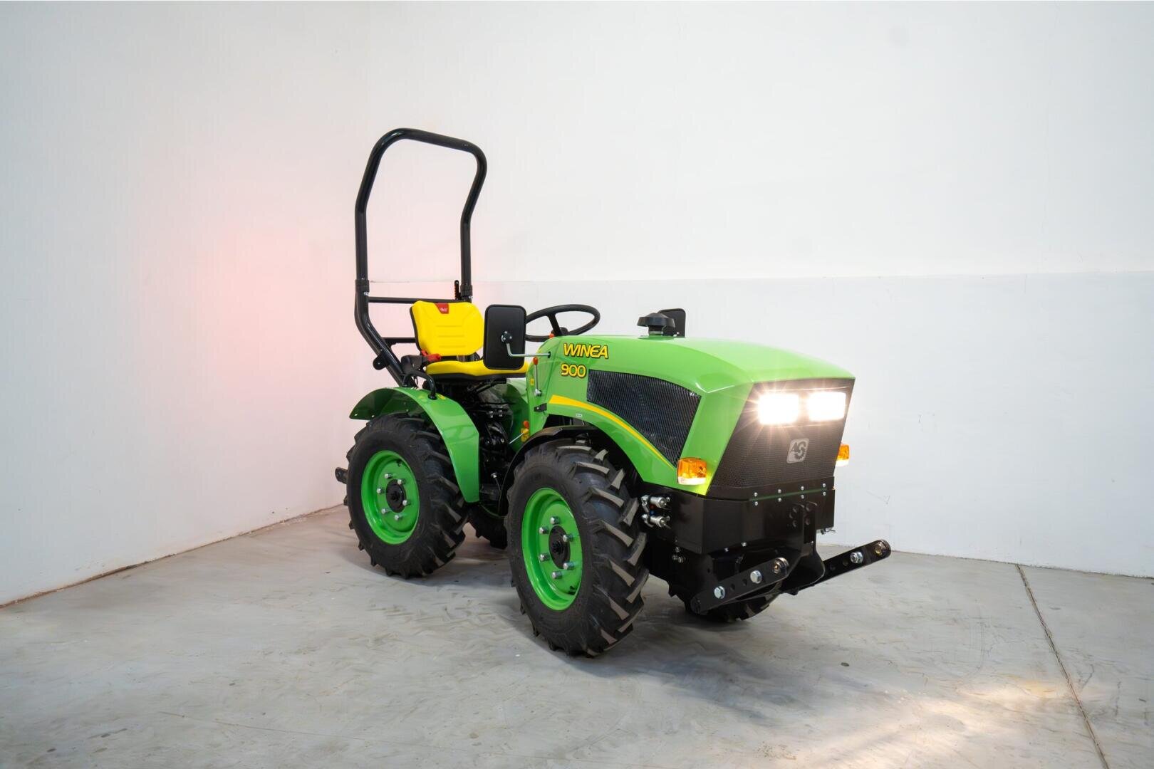 Ein grün-gelber Kleintraktor mit großen schwarzen Reifen und eingeschalteten Scheinwerfern, der in einem Innenraum vor einer schlichten weißen Wand geparkt ist.
