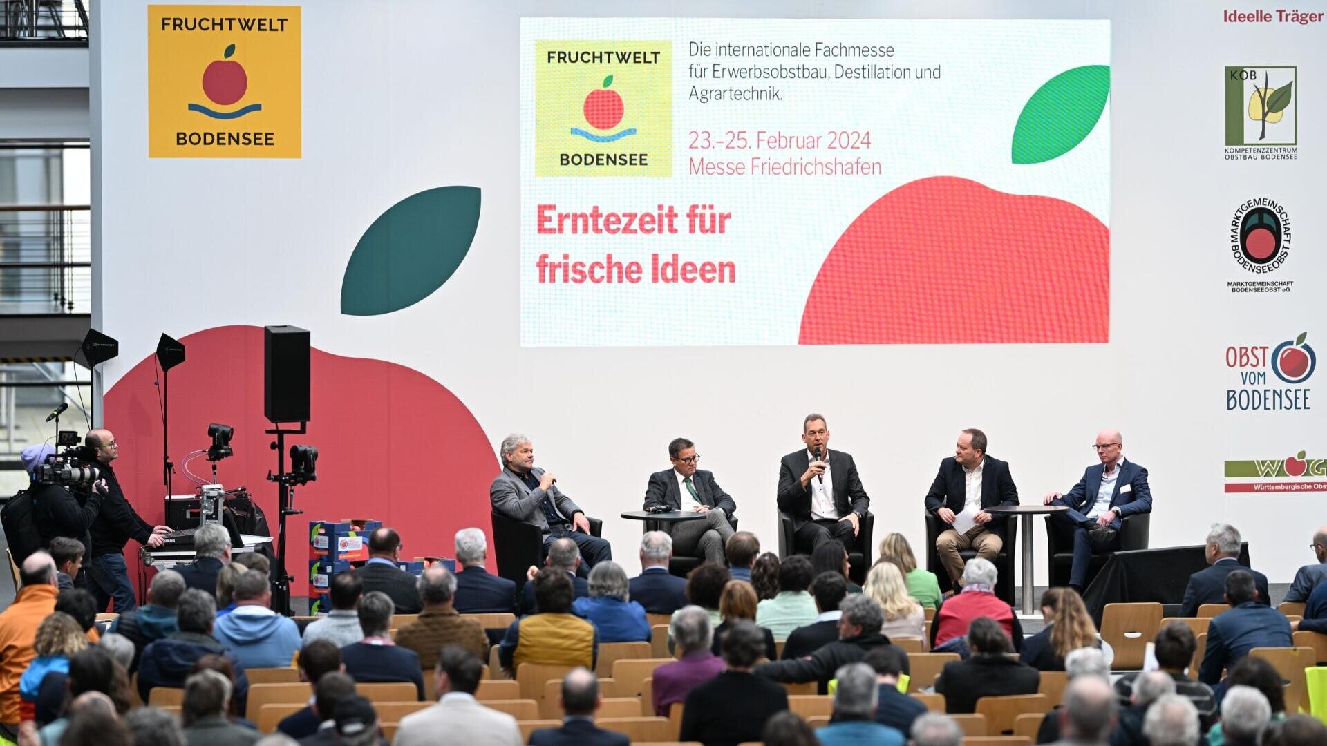 Eine Gruppe von fünf Männern sitzt auf der Bühne vor dem Publikum der Fruchtwelt Bodensee. Auf einem großen Hintergrund sind Veranstaltungshinweise und eine rote Apfelgrafik zu sehen. Ein Mann spricht, während die anderen zuhören.