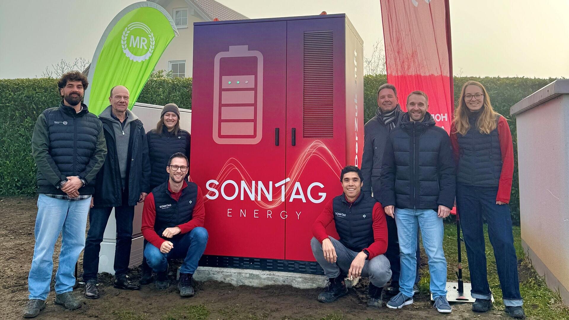 Acht Personen stehen lächelnd um einen großen roten Energiespeicher mit der Aufschrift SONNTAG ENERGY und einem Batteriesymbol. Im Hintergrund sind Grünflächen, Banner und ein Gebäude zu sehen.