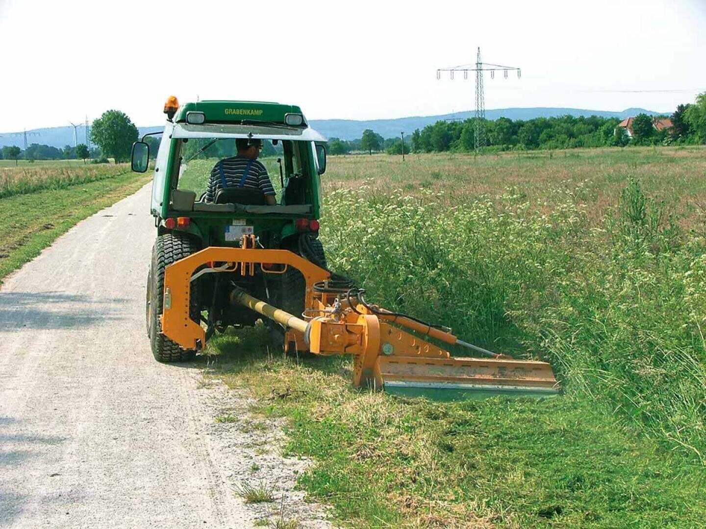 Eine Person fährt einen grünen Traktor mit einem gelben Mähwerk und mäht Gras am Rande einer ländlichen Schotterstraße, die von Feldern und Vegetation gesäumt ist.