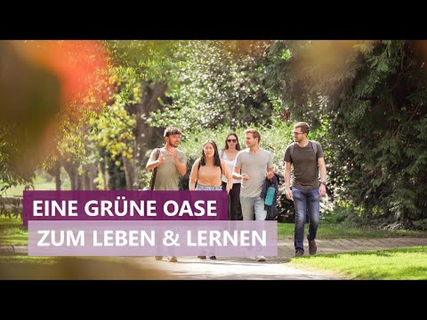 A group of five young adults go for a walk together on a sunny path in the countryside. The text in German reads EINE GRÜNE OASE ZUM LEBEN & LERNEN.