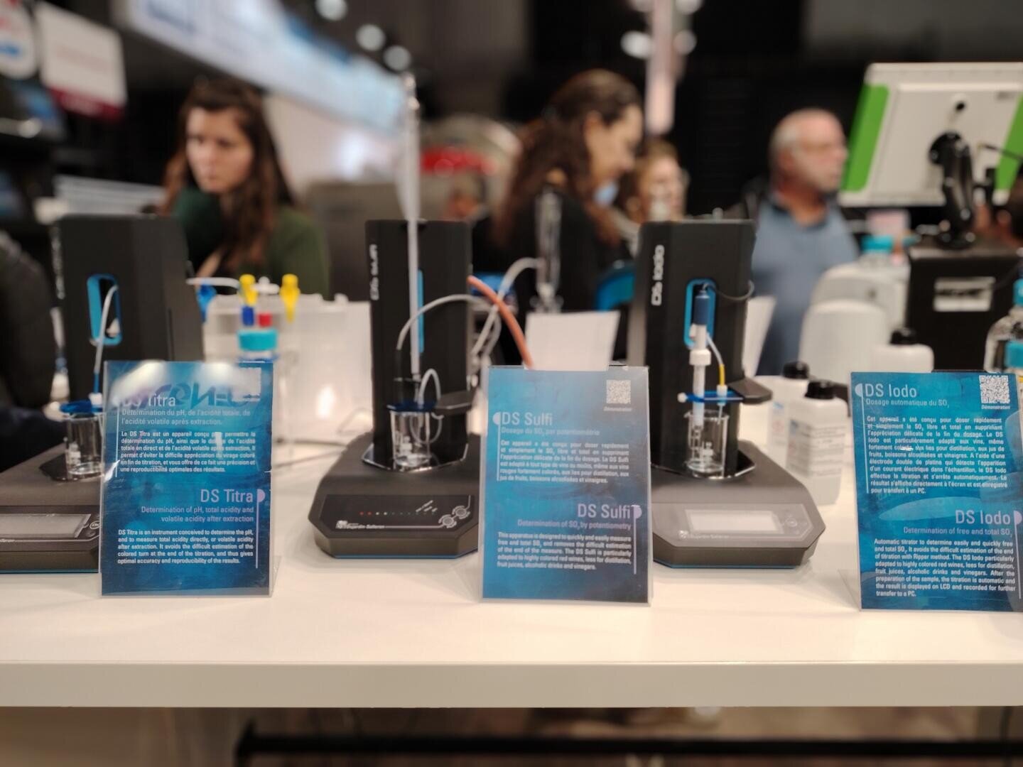 Nahaufnahme von wissenschaftlichen Geräten und Schautafeln auf einem Labortisch, mit Menschen, die im unscharfen Hintergrund stehen und sich unterhalten. Auf den Schautafeln steht blauer Informationstext über verschiedene wissenschaftliche Instrumente.