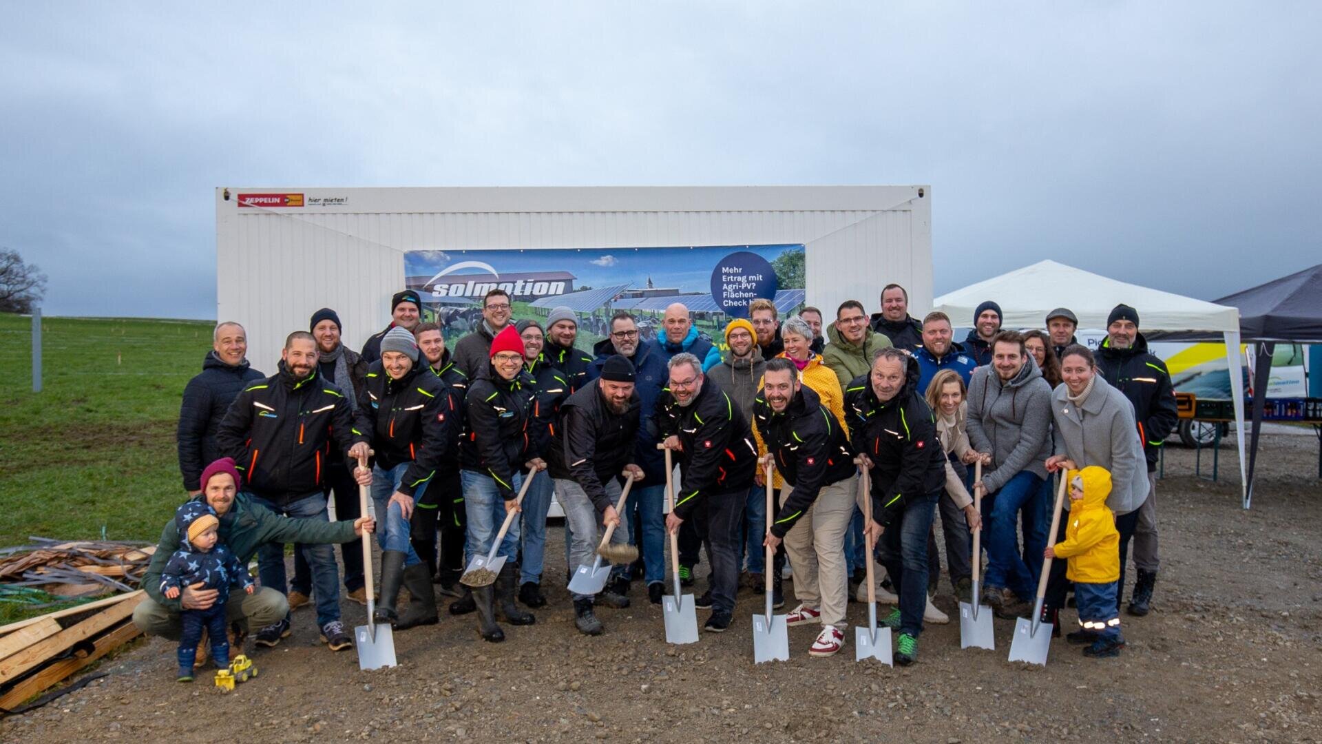 A large group of people, some with shovels in their hands, pose and smile outdoors in front of a white container labeled "solmotion". It seems to be a groundbreaking event on a cloudy day.