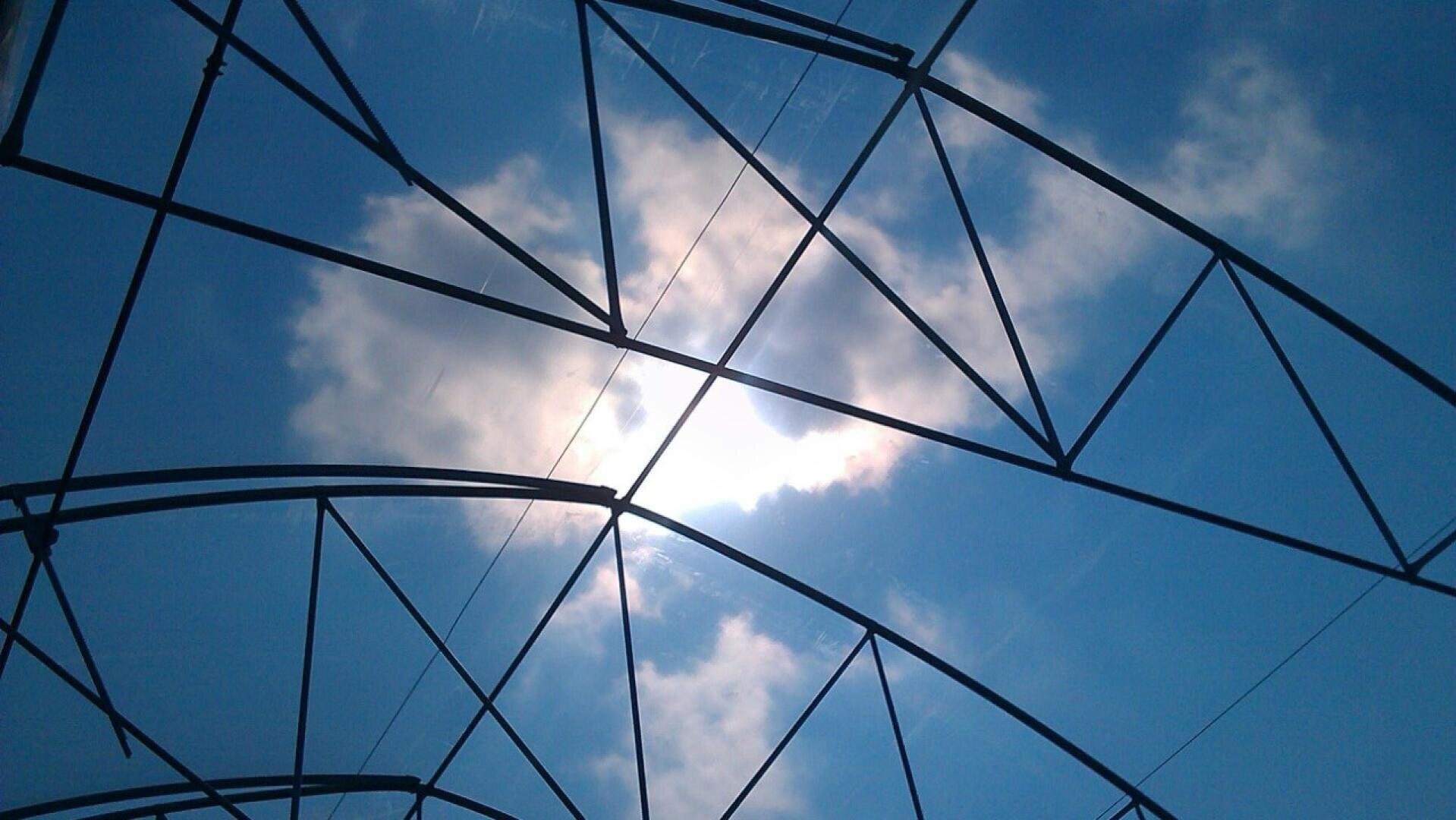 Metallgerüst eines Daches oder einer Struktur, das sich gegen einen hellen Himmel mit vereinzelten Wolken und durch die Lücken scheinendem Sonnenlicht abhebt.