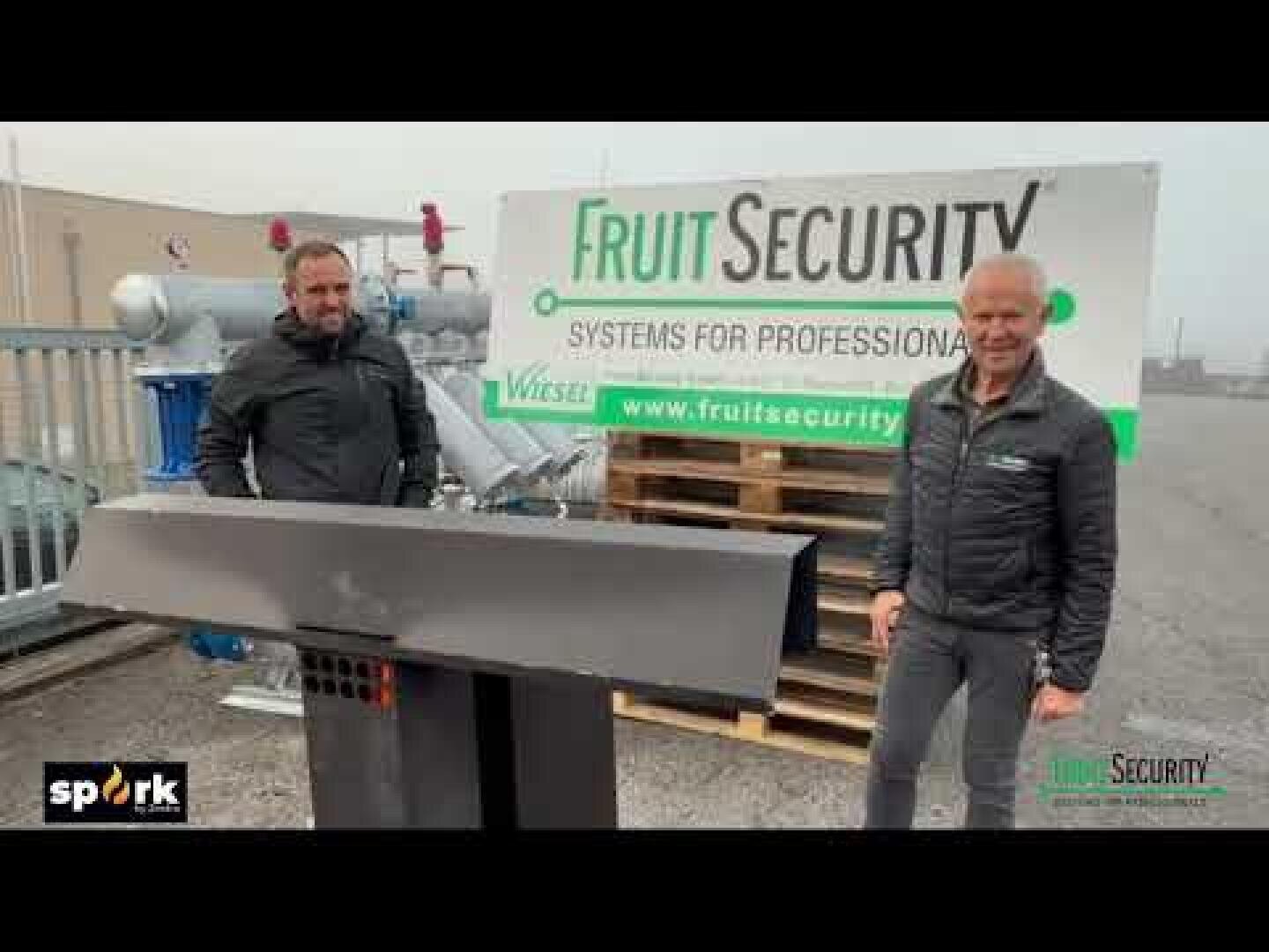 Zwei Männer stehen im Freien neben Industrieanlagen, vor einem Banner mit der Aufschrift Fruit Security: Systeme für Fachleute. Im Hintergrund sind Paletten und Maschinen zu sehen. Die Logos Spork und Fruit Security erscheinen auf dem Bild.