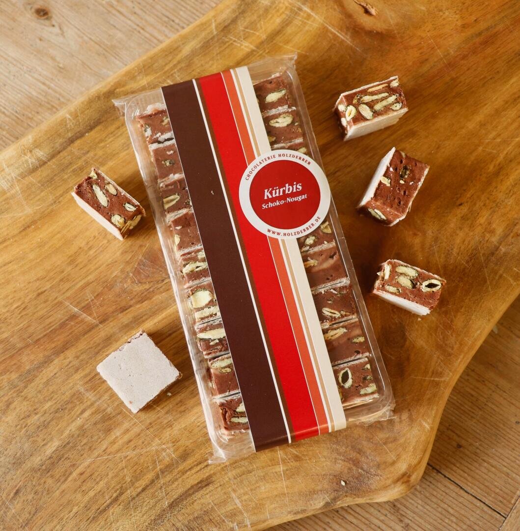 Auf einem Holzbrett liegt eine Packung Kürbis Schoko-Nougat-Konfekt, umgeben von mehreren ausgepackten Nougatstücken mit sichtbaren Nüssen und Schichten.