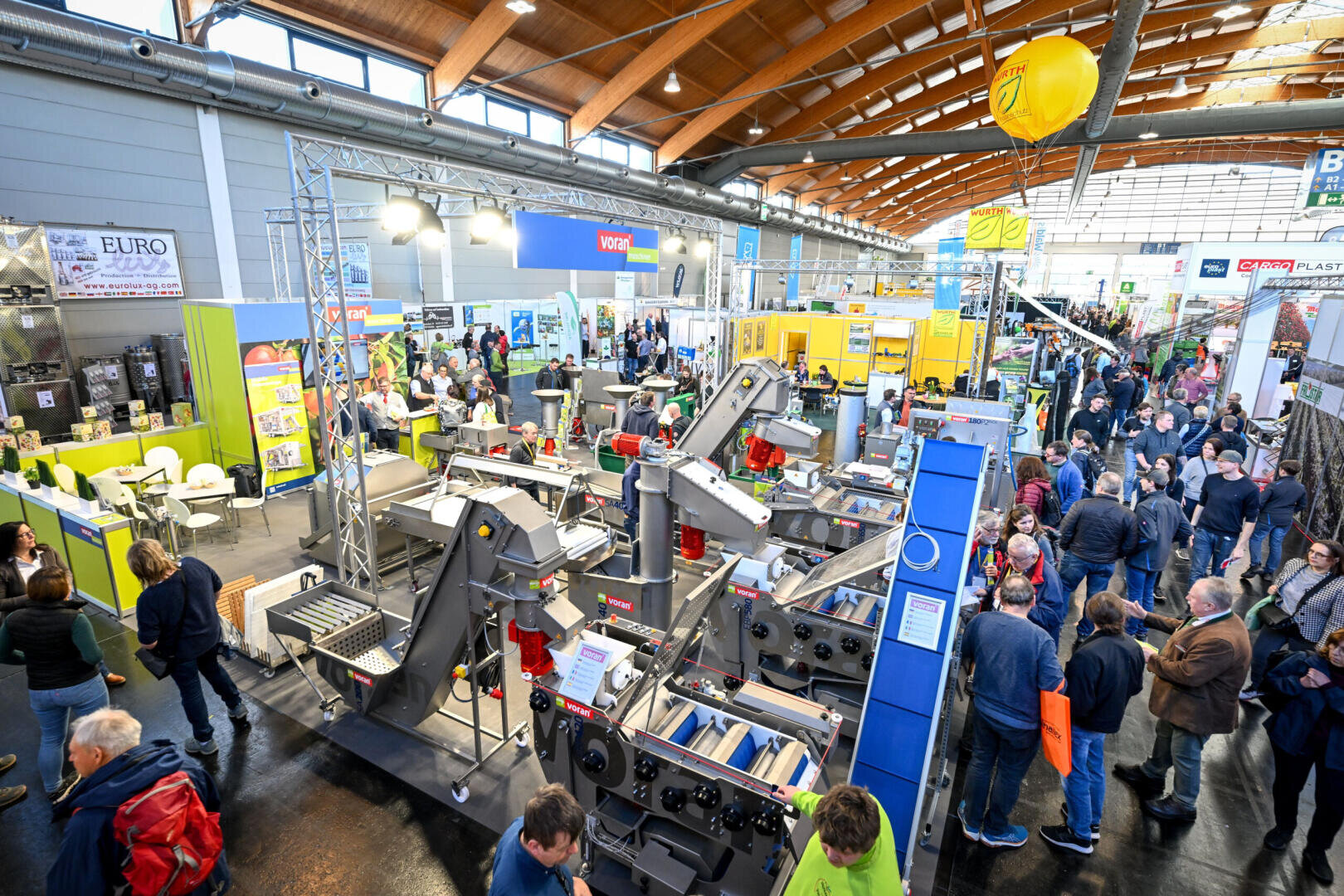Eine geschäftige Messe mit vielen Menschen, die zwischen Industriemaschinen und bunten Ausstellerständen in einer großen Halle mit einer hohen Holzdecke umherlaufen.