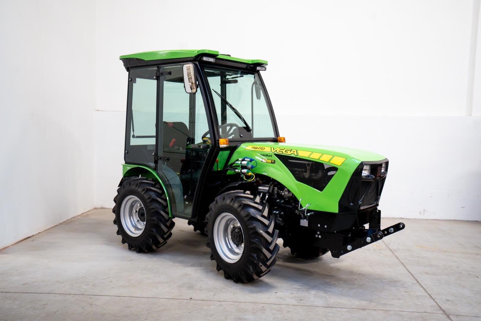 A green, modern agricultural tractor with a glass cab stands on a clean concrete floor in a hall in front of a white wall. The vehicle has large, robust tires and the lettering "Vega" on the side.