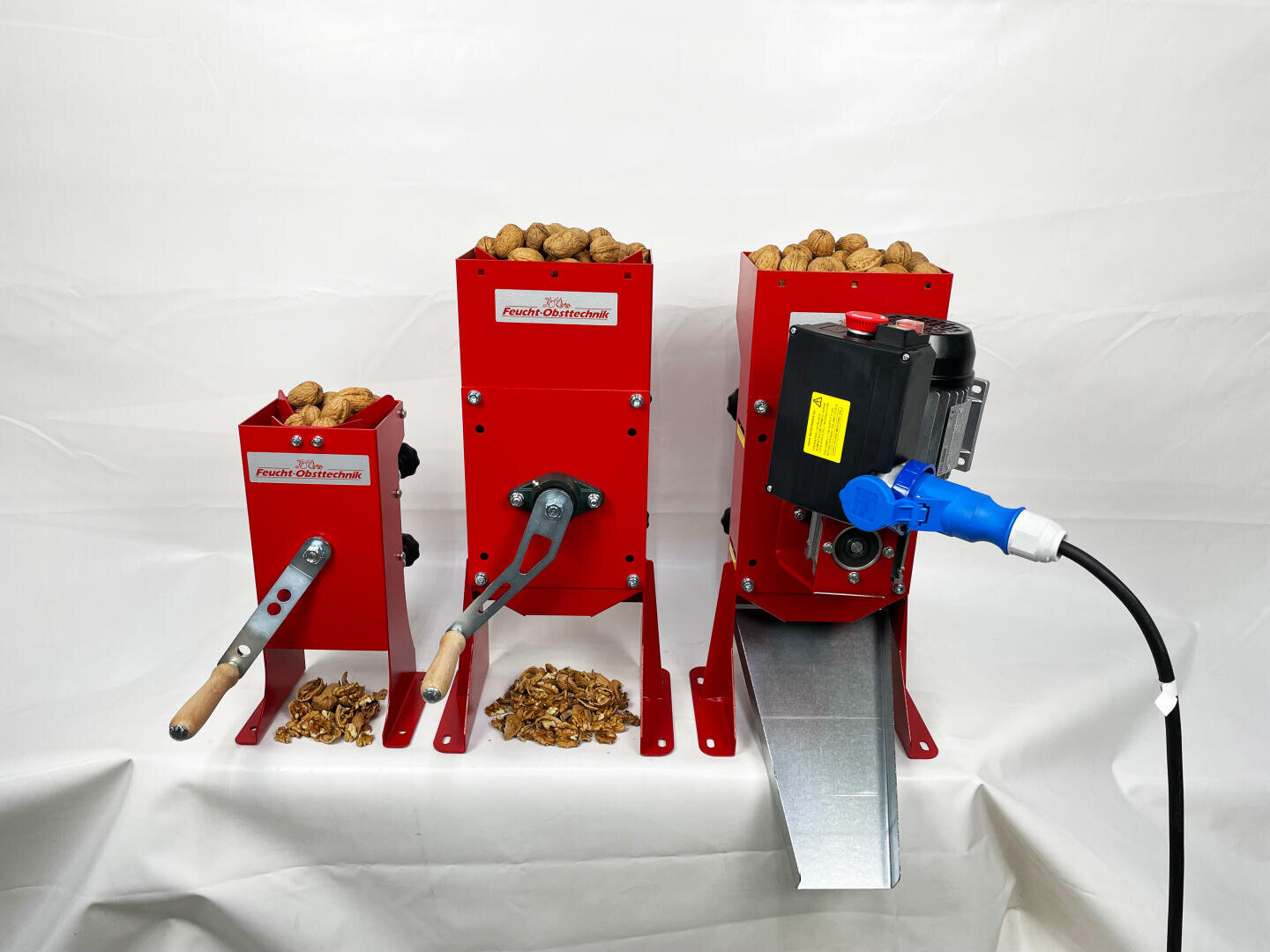 Three red nut cracking machines, each filled with walnuts. The left and center machines have manual cranks, while the right machine is electric and has a blue power plug and metal chute. Cracked walnut shells can be seen under each machine.