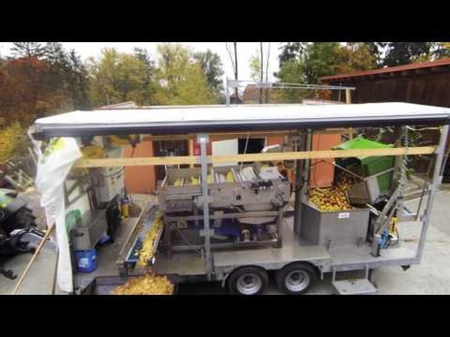 Eine mobile Obstverarbeitungsanlage auf einem Anhänger mit Förderbändern und Behältern, gefüllt mit gelben Früchten, möglicherweise Äpfeln, umgeben von Bäumen und Außenanlagen.