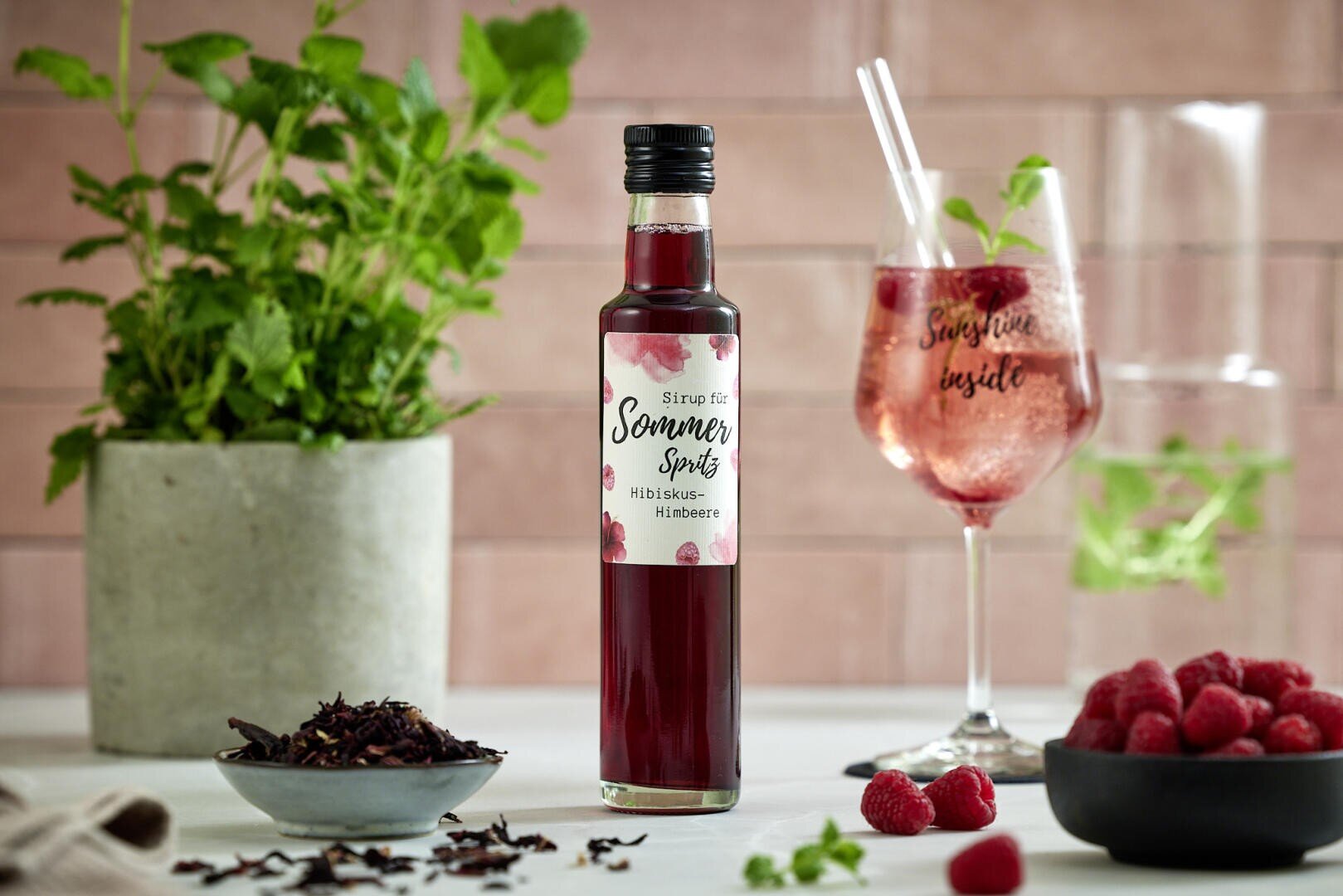 Eine Flasche Himbeersirup steht auf einem Tisch neben einem Glas rosa Getränk mit Eis und Minze, einer Schale mit Himbeeren, einer Schale mit getrocknetem Hibiskus und einer Topfpflanze mit Minze vor einem rosa gekachelten Hintergrund.