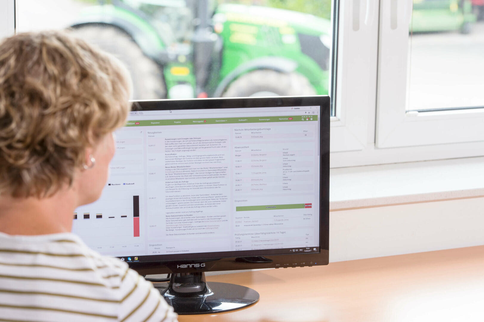 Eine Person mit kurzem, lockigem Haar sitzt an einem Schreibtisch und betrachtet Daten und Texte auf einem Computermonitor. Ein Fenster im Hintergrund zeigt einen verschwommenen grünen Traktor draußen.