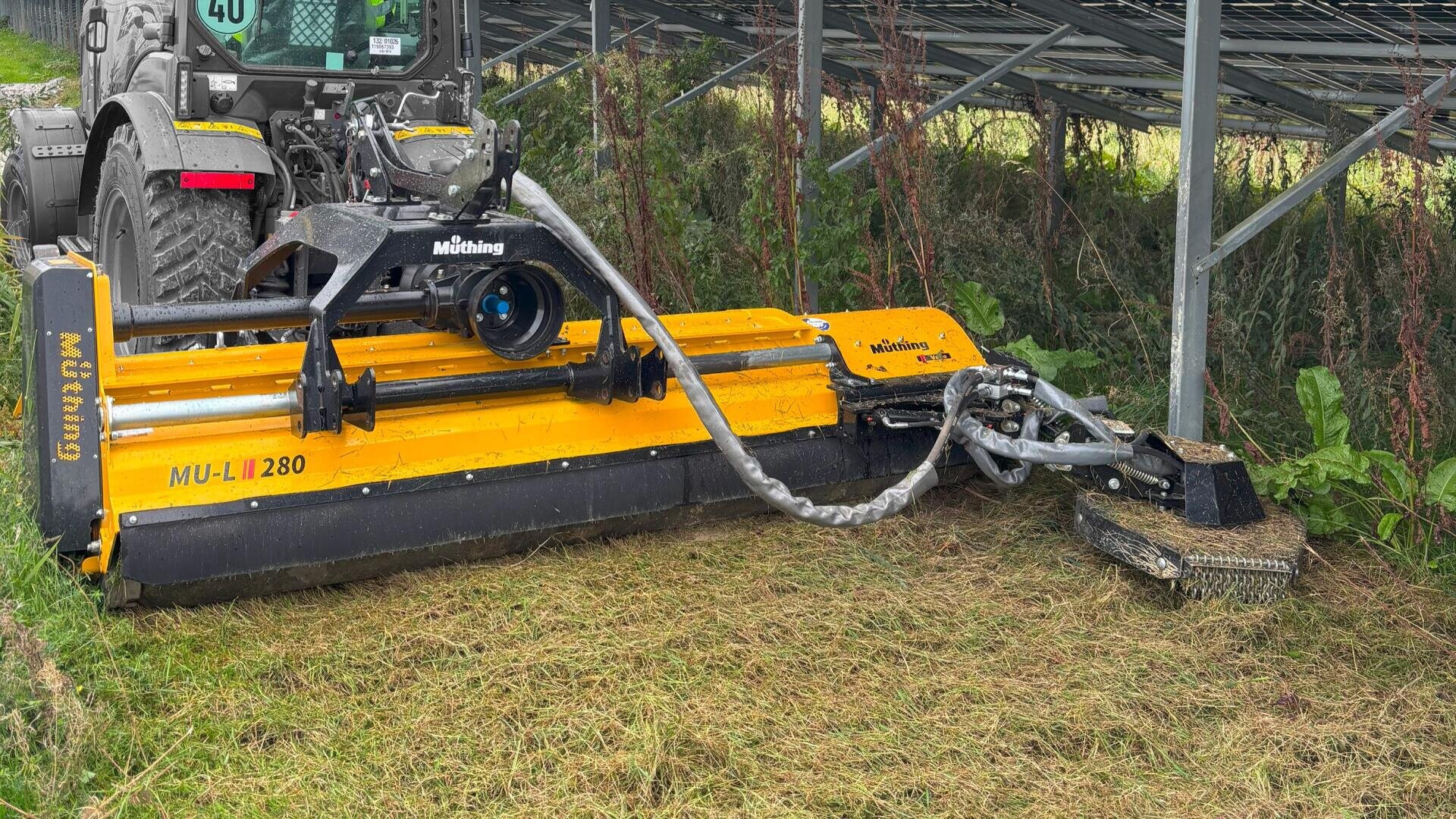 Ein gelber Mulchmäher, der an einen Traktor angehängt ist, mäht das Gras unter einer Solarzellenanlage, wobei das gemähte Gras und die grüne Vegetation rund um die Struktur sichtbar sind.