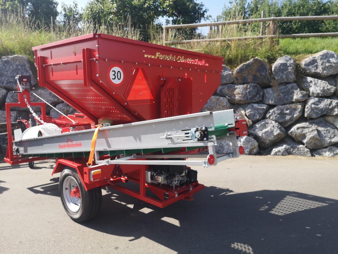 Eine rot-silberne Landmaschine steht auf einer gepflasterten Fläche in der Nähe einer Steinmauer und eines grasbewachsenen Hangs. An der Seite sind ein großer Trichter, ein Förderband, Räder und die Aufschrift Fliegl Obsttechnik zu sehen.