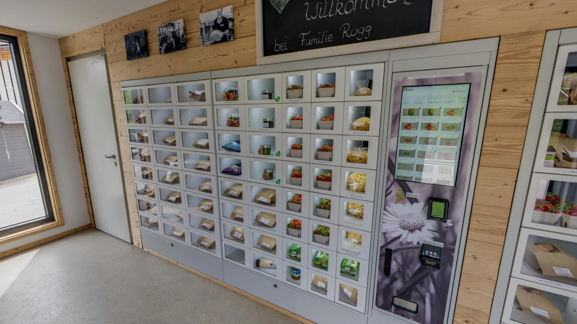 Ein an der Wand montierter Automat mit mehreren Fächern, die frische Produkte wie Erdbeeren und Eier enthalten, neben einem Bedienfeld in einem holzgetäfelten Raum.