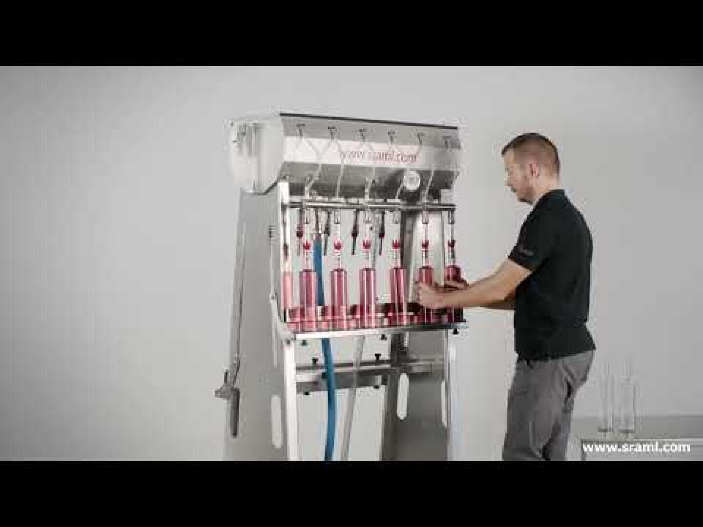 A man operates a semi-automatic bottle filling machine and fills several glass bottles with liquid. The machine has several filling nozzles and stands in front of a plain, light-colored wall.