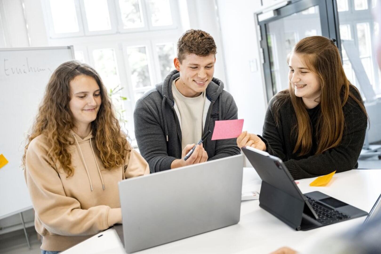 Drei junge Erwachsene sitzen an einem Tisch mit Laptops, lächeln und arbeiten zusammen. Einer hält einen rosafarbenen Zettel in der Hand, während ein anderer darauf schreibt. Sie scheinen sich in einem hellen, modernen Büro oder Klassenzimmer zu befinden.