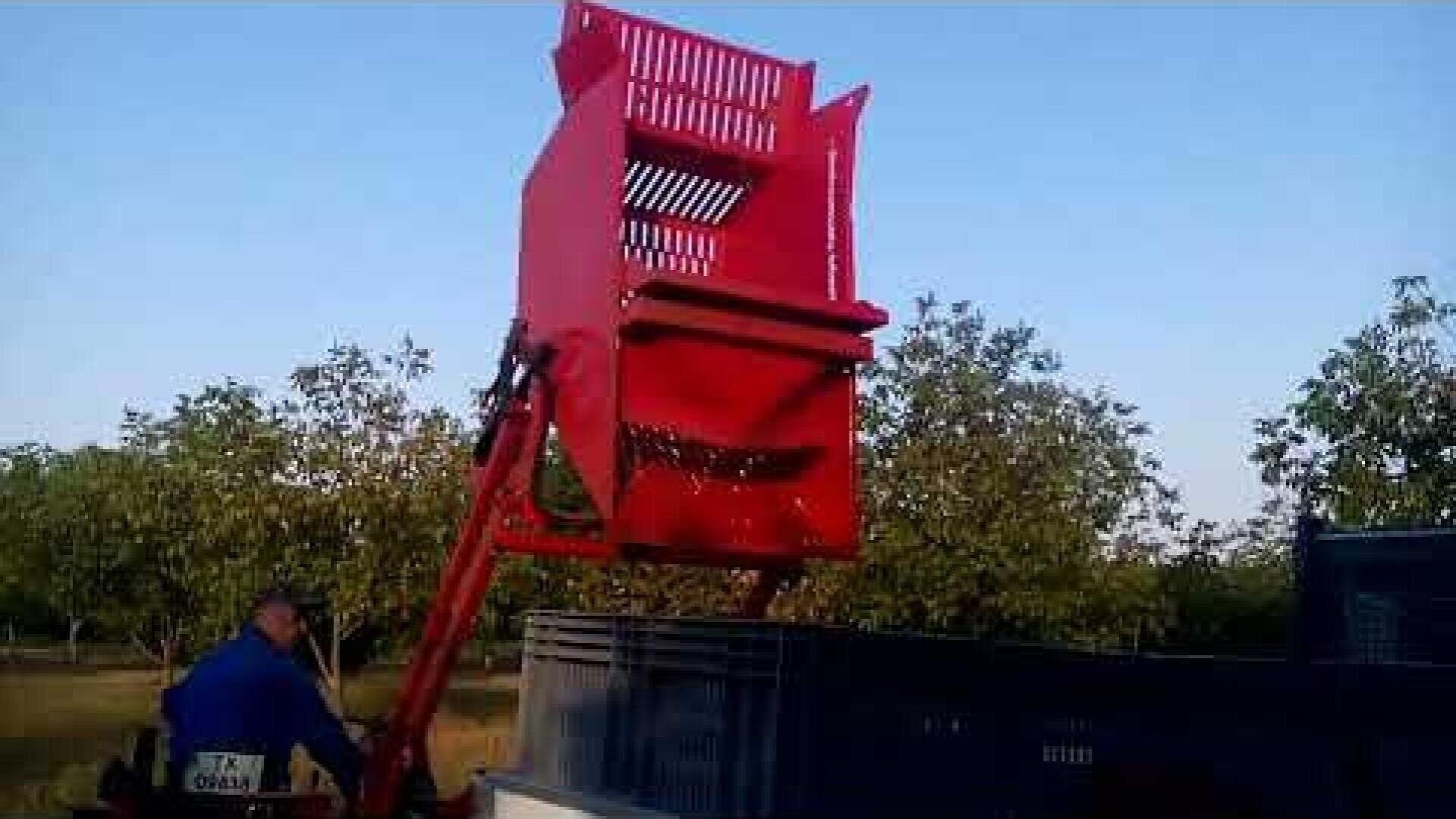 A red mechanical harvester lifts a container of nuts and empties it into a large bin, while a person operates the machine in an orchard. Trees and a clear sky can be seen in the background.