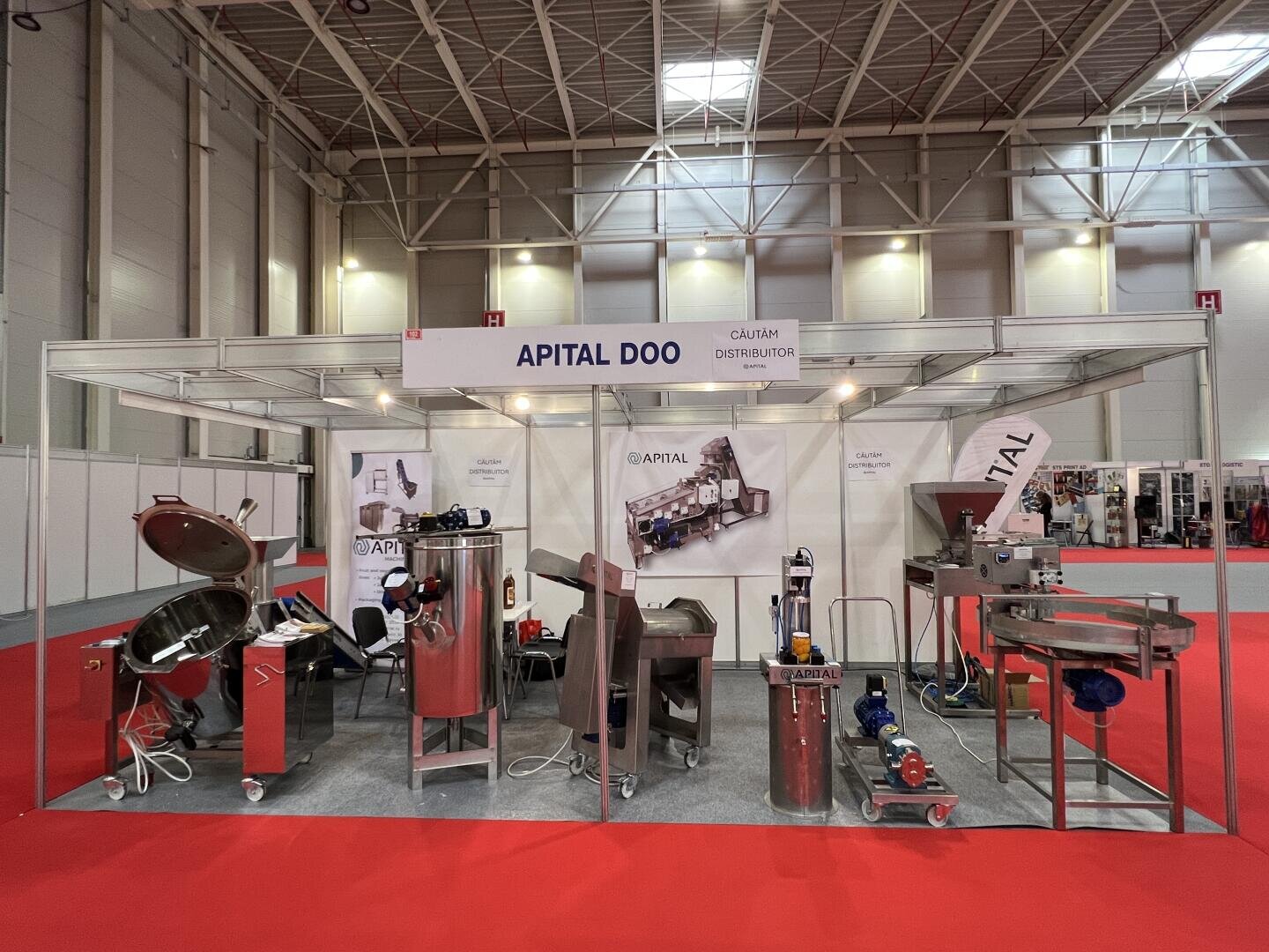 Ein Messestand von APITAL DOO mit industriellen Maschinen und Geräten aus rostfreiem Stahl auf einem grauen Boden mit rotem Teppich. Große Banner und Informationsplakate sind hinter den Geräten zu sehen.
