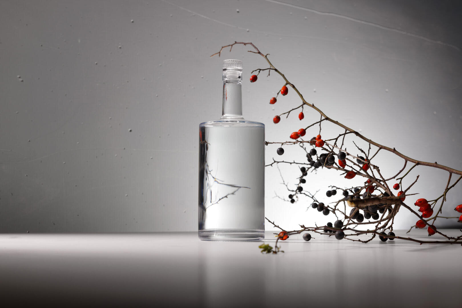 A clear glass bottle filled with transparent liquid stands on a smooth surface next to a dry branch with red and black berries against a gray background with dramatic lighting.