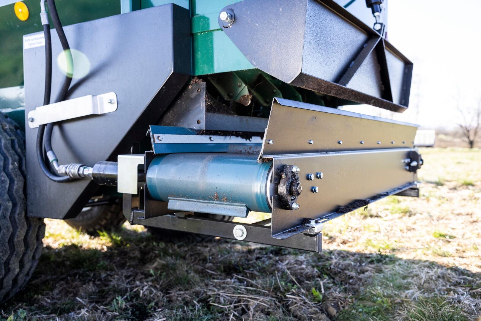 Close-up of a mechanical part on a green industrial machine with a blue hydraulic cylinder and metal parts, with grass and sunlight in the background.