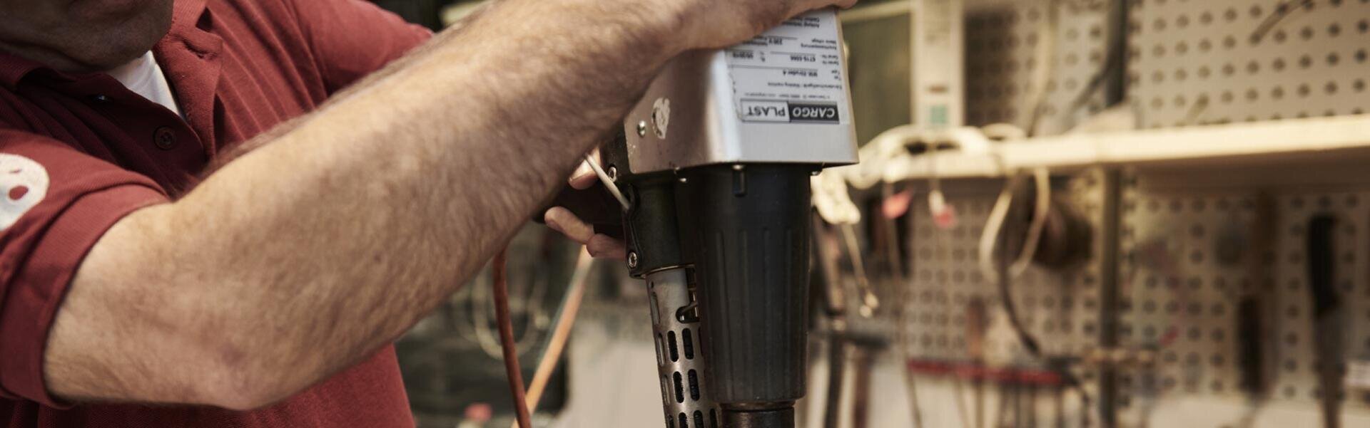 A person in a red shirt uses a hand-held power tool in a workshop, while various tools hang on a pegboard in the background.
