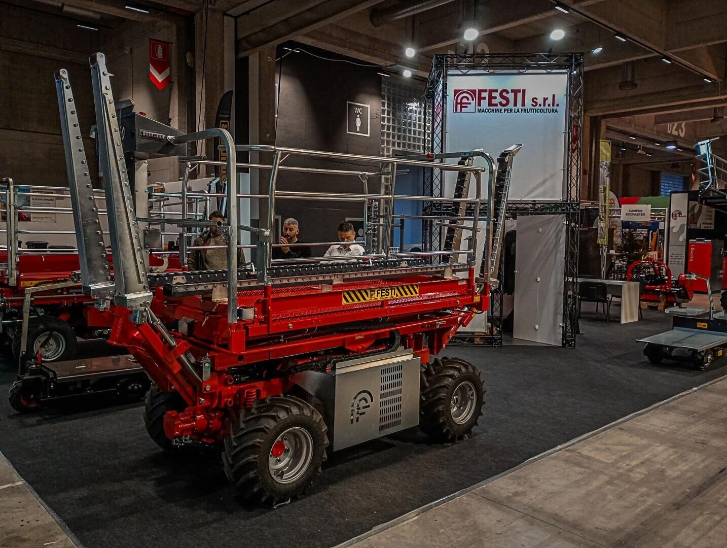 A red industrial lifting machine is displayed at an indoor expo booth for FESTI S.r.l., with people behind a counter and exhibition banners in the background. The setting is a large, well-lit hall.