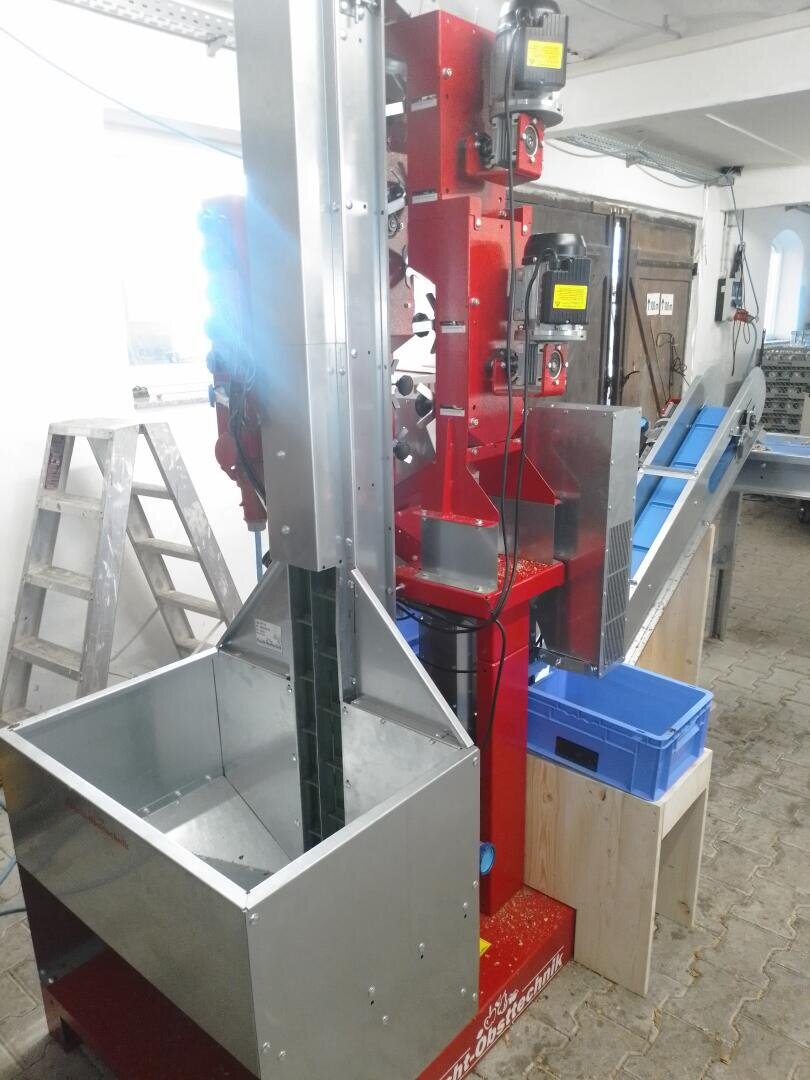 Eine vertikale rot-silberne Industriemaschine mit Motoren und einem Förderband steht in einer Werkstatt mit einer Metallleiter und blauen Kunststoffbehältern in der Nähe. Die Maschine scheint zum Sortieren oder Transportieren von Kleinteilen gedacht zu sein.