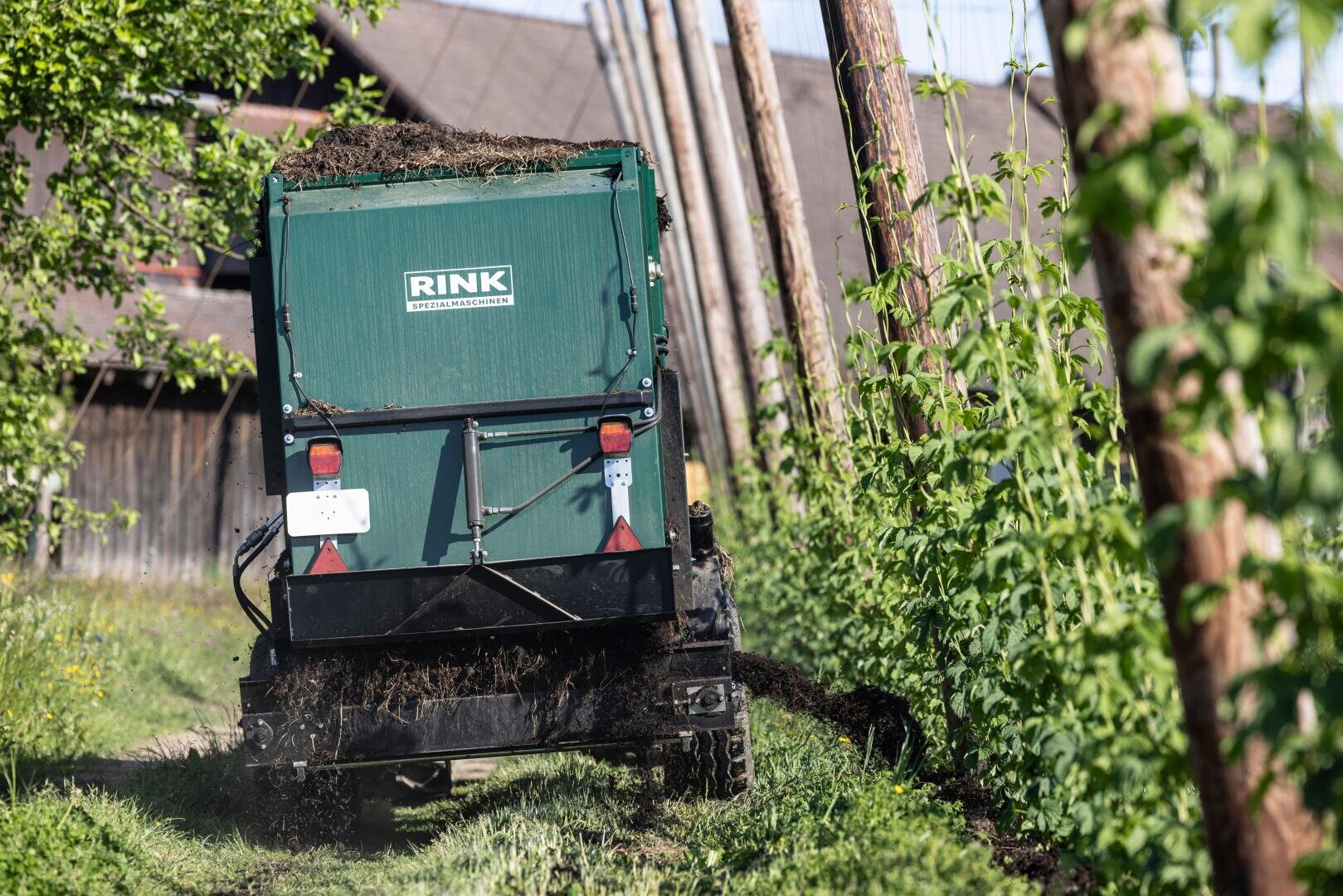 A green RINK agricultural machine spreads compost or manure on a field lined with tall plants (right). In the background, wooden stakes and a rustic building can be seen in the bright sunlight.
