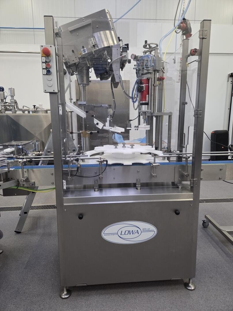 Eine industrielle Flaschenabfüll- und Verschließmaschine aus Edelstahl mit Bedientasten, Förderband und mechanischen Armen in einer Fabrikumgebung. Die Maschine ist mit LOWA beschriftet.