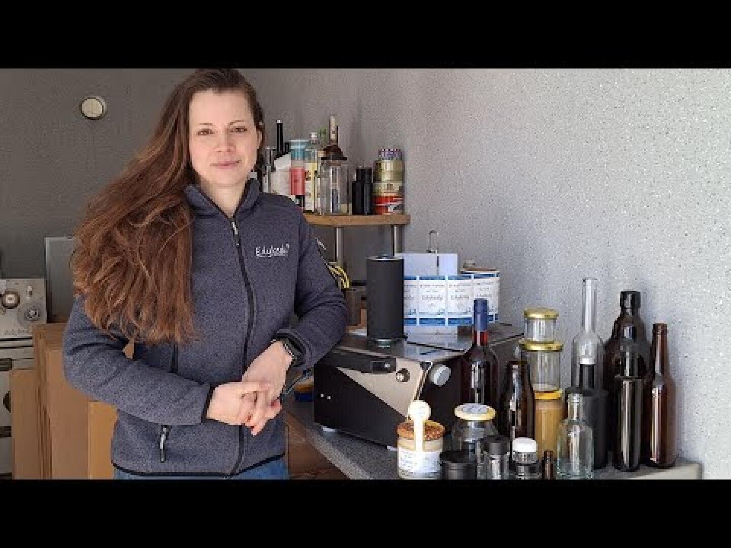 A woman with long brown hair and a gray jacket stands in a room with light gray walls next to a table with various bottles, glasses and utensils.