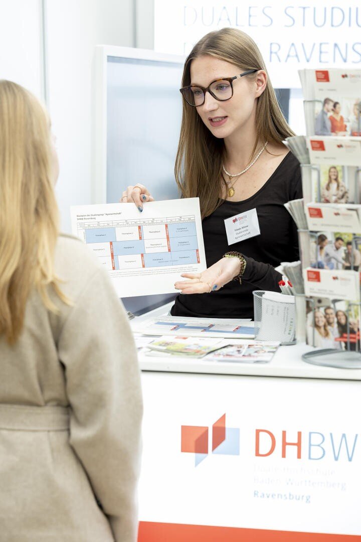 Eine Frau an einem Stand der DHBW Ravensburg erklärt einer anderen Frau ein Informationsblatt. Am Stand liegen Broschüren aus, und die beiden Frauen scheinen bei einer Bildungsveranstaltung ins Gespräch zu kommen.
