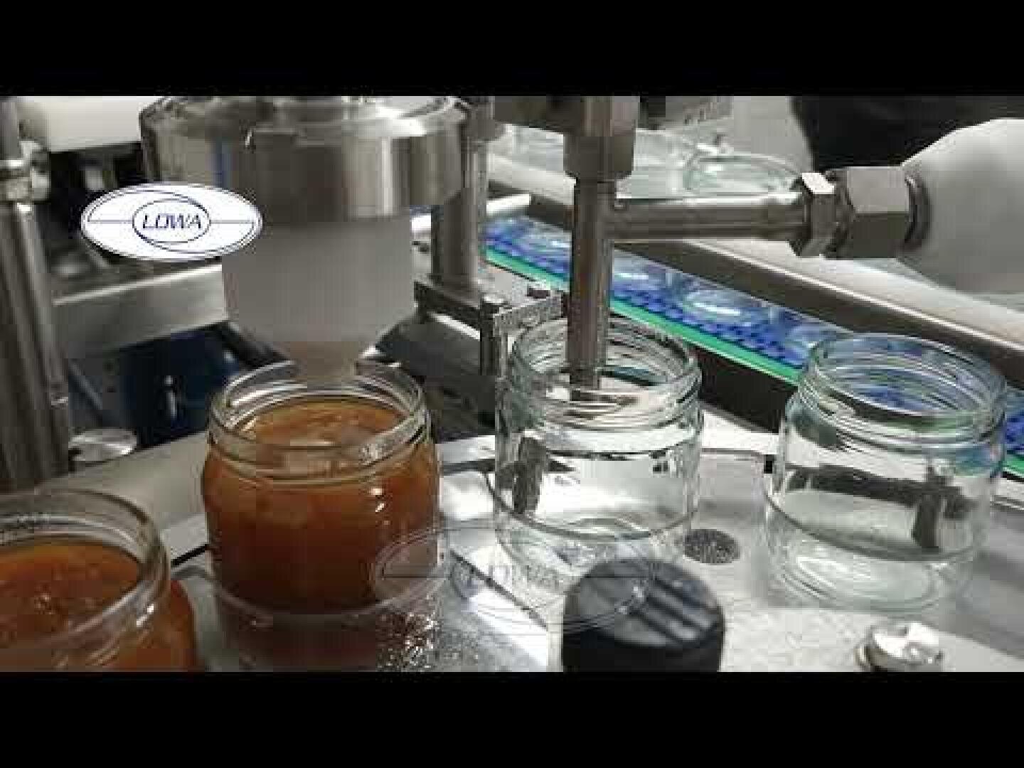 Nahaufnahme einer Lebensmittelverarbeitungsmaschine, die auf einem Fließband Gläser mit einer braunen, marmeladenartigen Substanz füllt. Die Gläser bewegen sich in einer kreisförmigen Bewegung, und einige werden gefüllt, während andere leer sind.