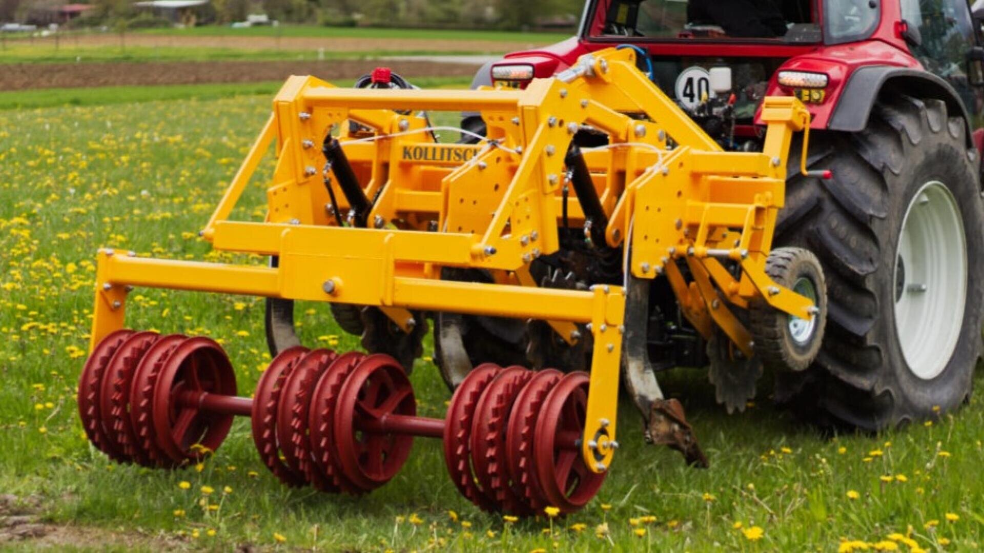 A red tractor is hitched to a large yellow agricultural implement with red rollers, parked in a grassy field with scattered yellow flowers.