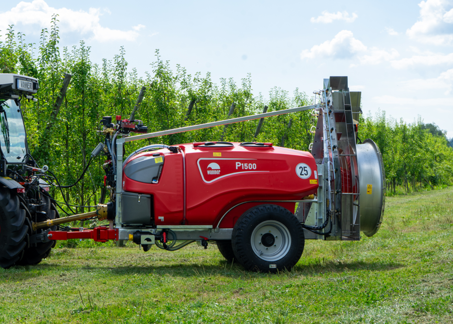 Eine rote Feldspritze mit der Aufschrift P1500 ist an einem Traktor in einem Obstgarten mit grünen Baumreihen unter einem teilweise bewölkten Himmel angebracht.