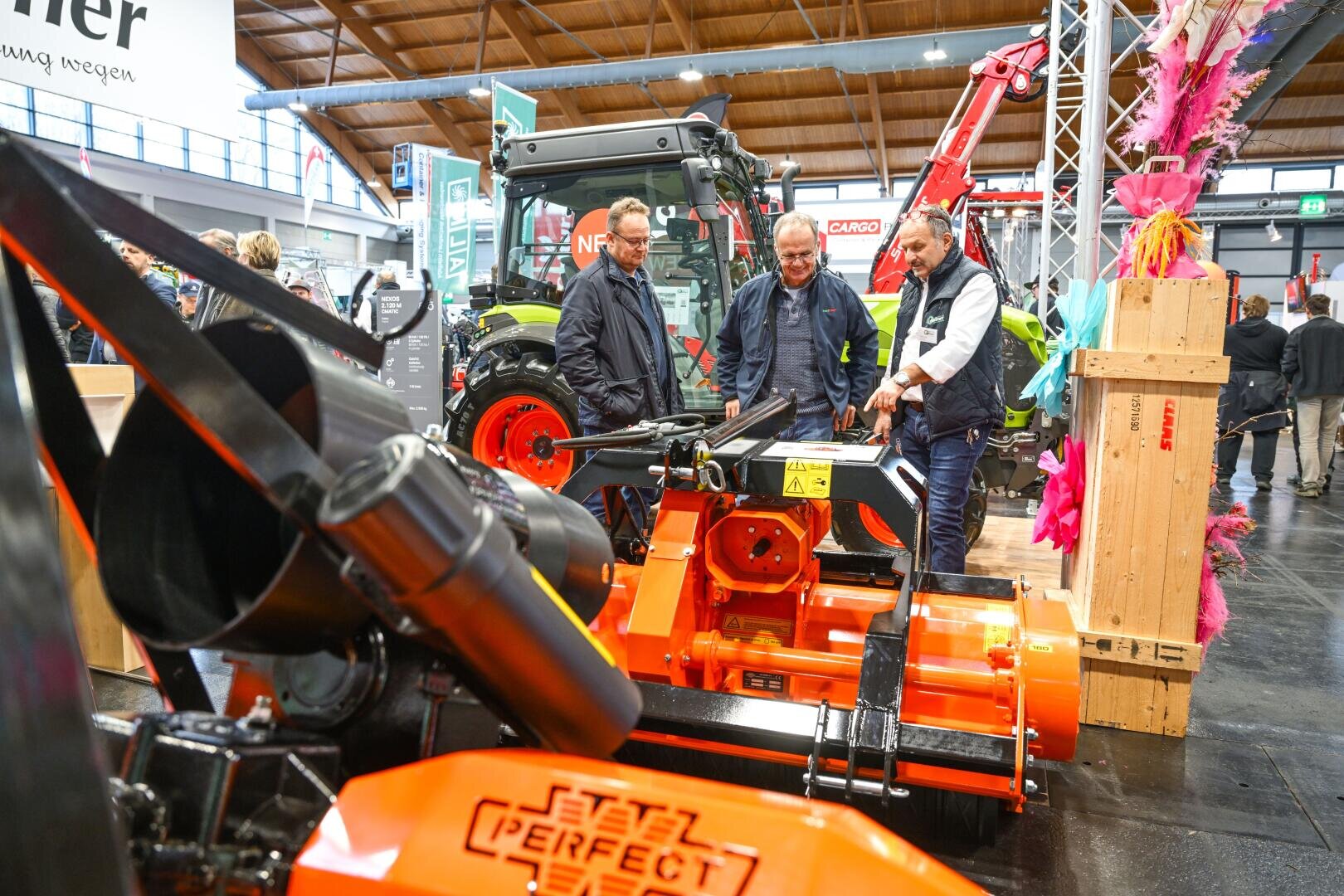 Three men stand at an indoor machinery exhibition and discuss a large orange agricultural machine. Tractors and other agricultural equipment can be seen in the background, while wooden crates and colorful decorations stand nearby.