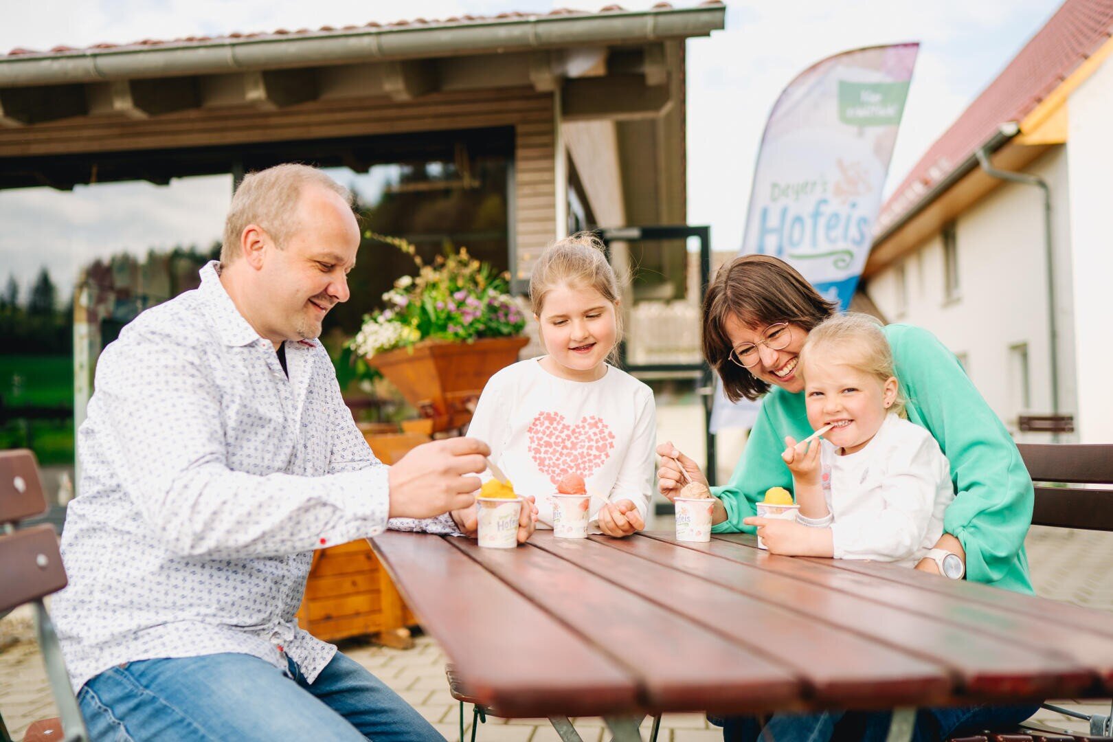 Eine lächelnde vierköpfige Familie sitzt an einem Holztisch im Freien und genießt gemeinsam ein Eis. Die Eltern und zwei junge Mädchen lachen und unterhalten sich, während im Hintergrund Gebäude und Grünflächen zu sehen sind.